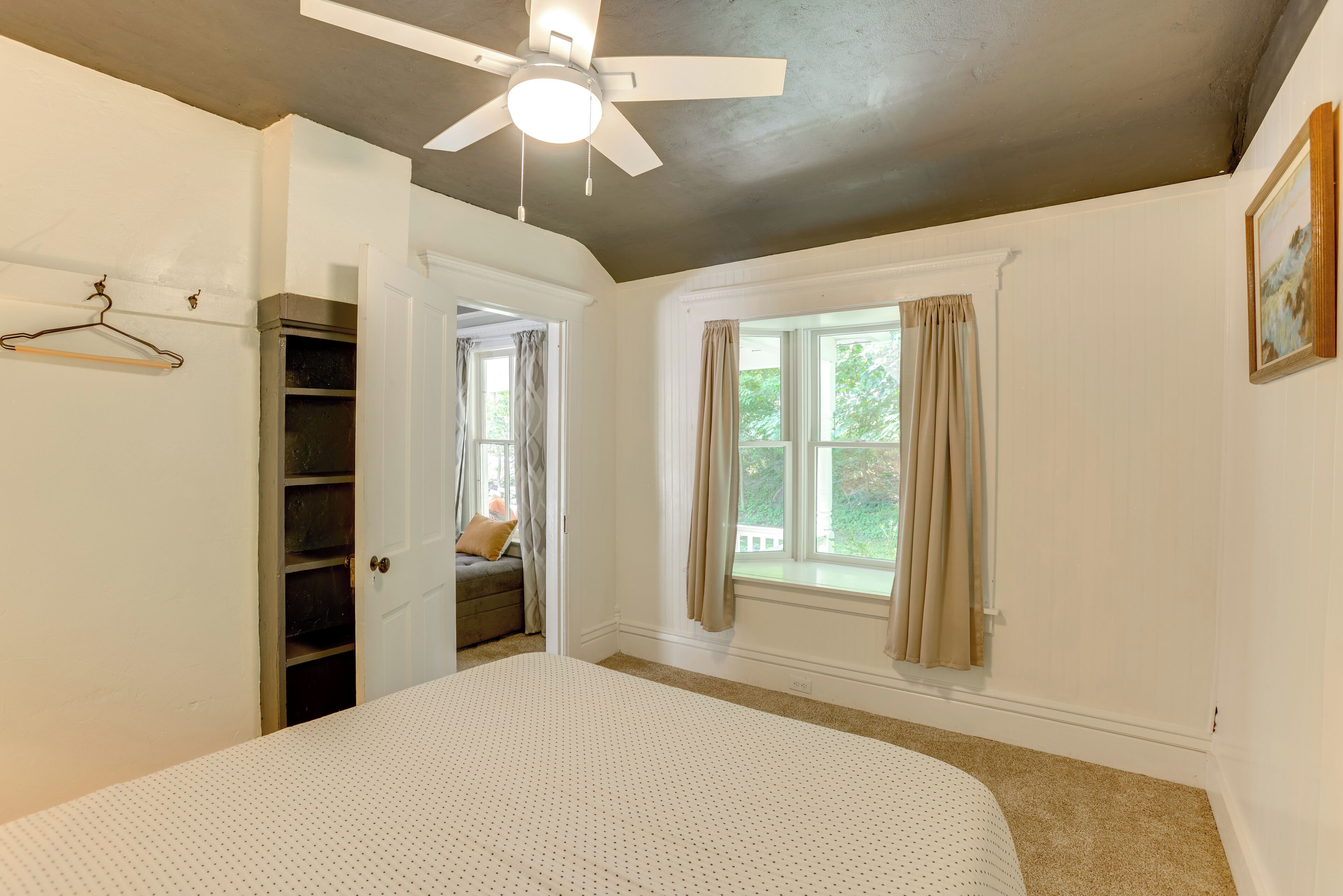 Quarto com cama, ventilador de teto, janela com cortinas e porta para outro cômodo.