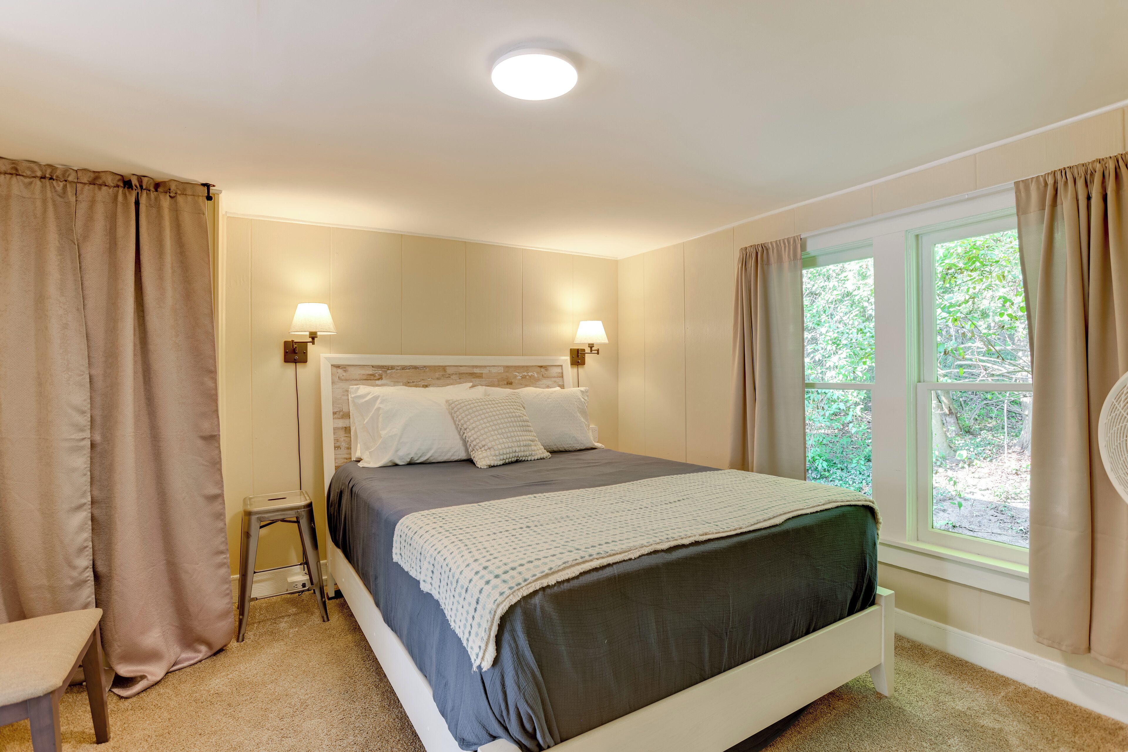 A bedroom with a bed, two bedside lamps, a window with curtains, a small stool, and a chair.