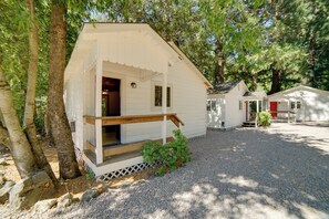A white cabin with a porch, surrounded by trees and gravel. - Golf & Hike: Cobb Mountain Cabin in Scenic Forest! (Cobb)
