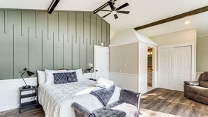 A bedroom with a bed, bedside tables, a ceiling fan, a bench, and a door leading to another room. - Hot Tub + Game Room! Lakefront Gem in Pocono Mtns (Tobyhanna)