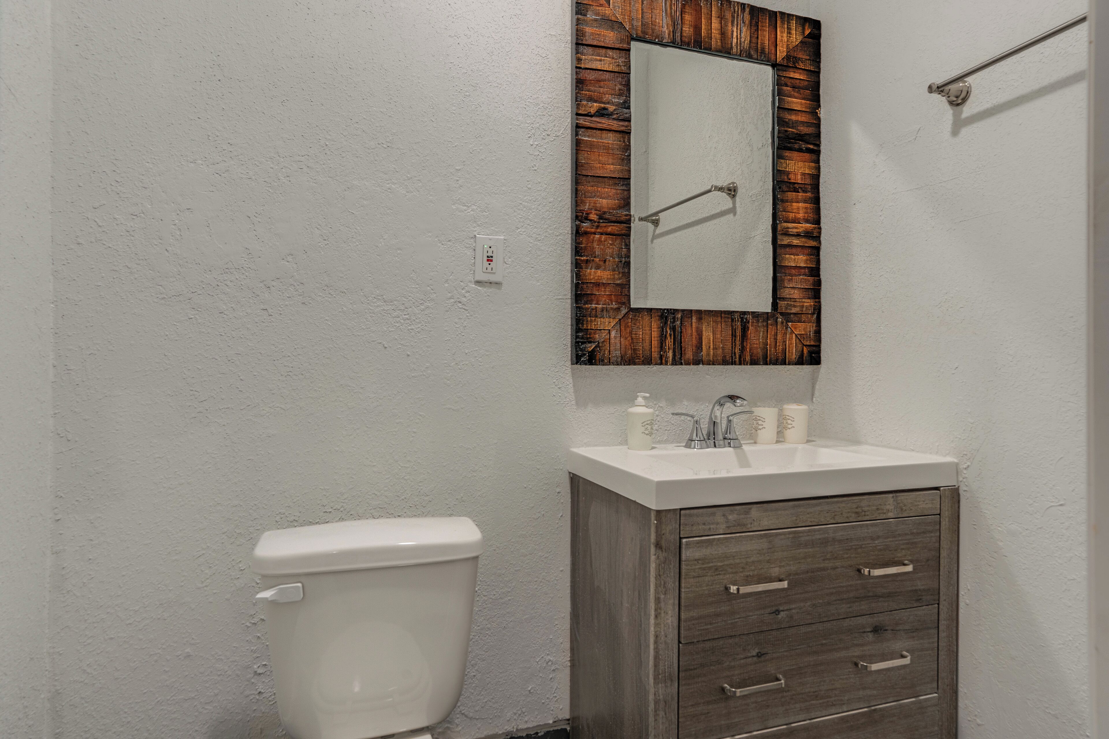Un baño con inodoro blanco, tocador de madera con cajones, espejo con marco de madera y lavabo blanco.
