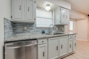 Una cocina con armarios blancos, encimera de granito, lavavajillas de acero inoxidable y una ventana con persianas.