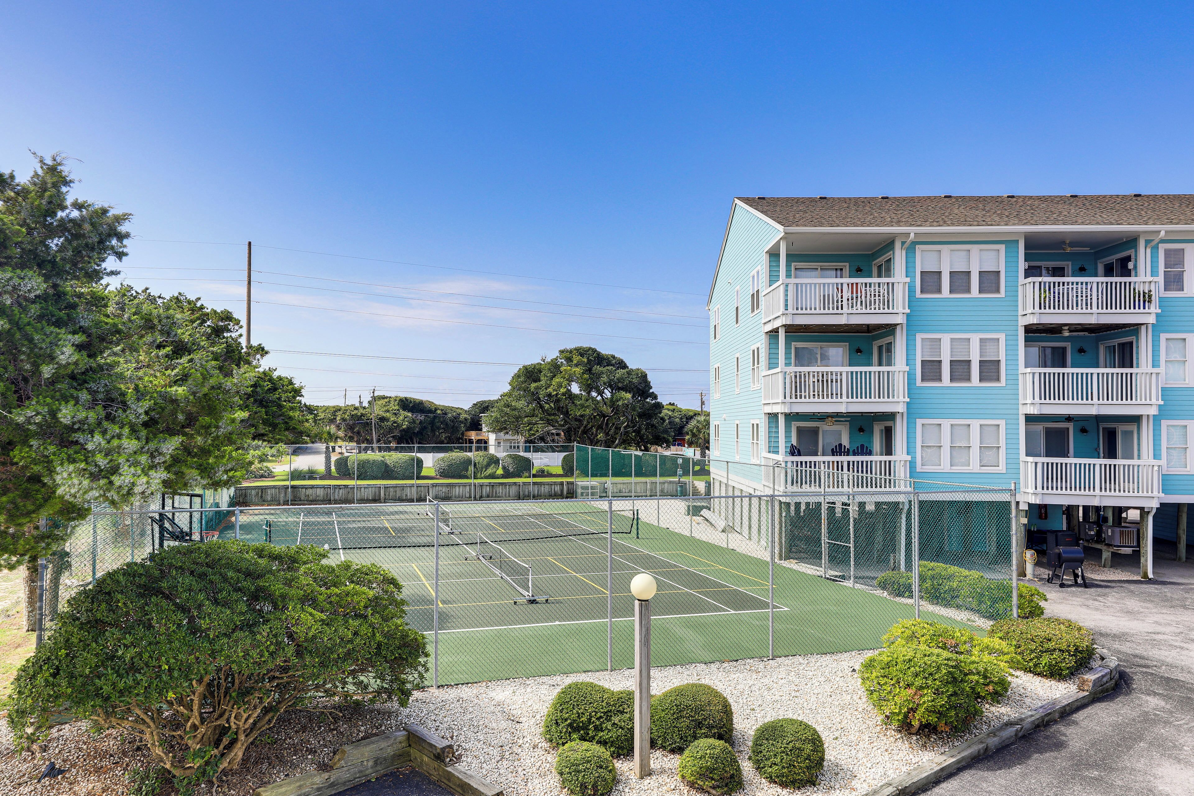 Ein von einem Zaun umgebener Tennisplatz, im Hintergrund ein Gebäude mit Balkonen und ein klarer, blauer Himmel.