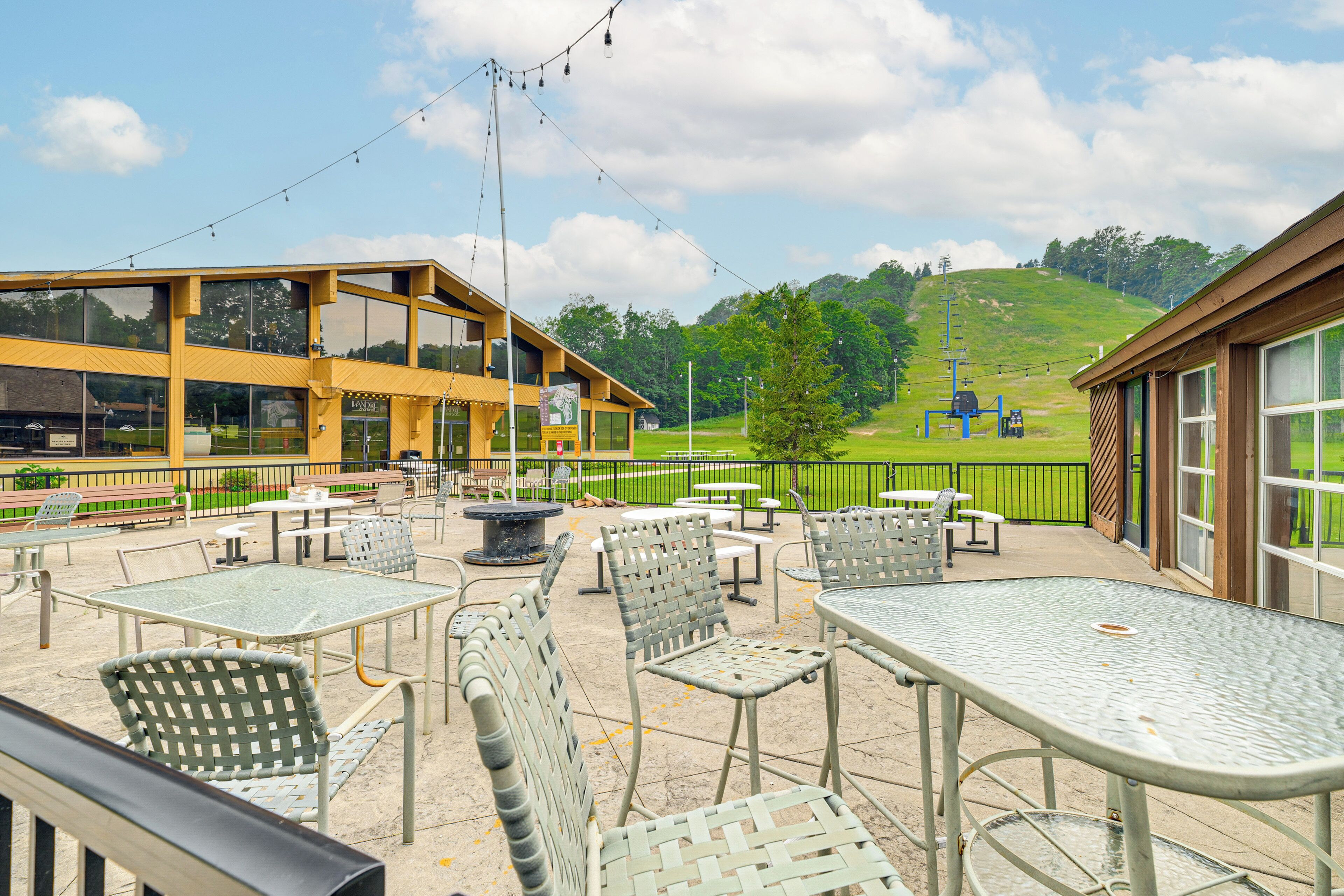 An outdoor patio area with tables and chairs, a building with large windows, and a view of a hill with a ski lift in the background.