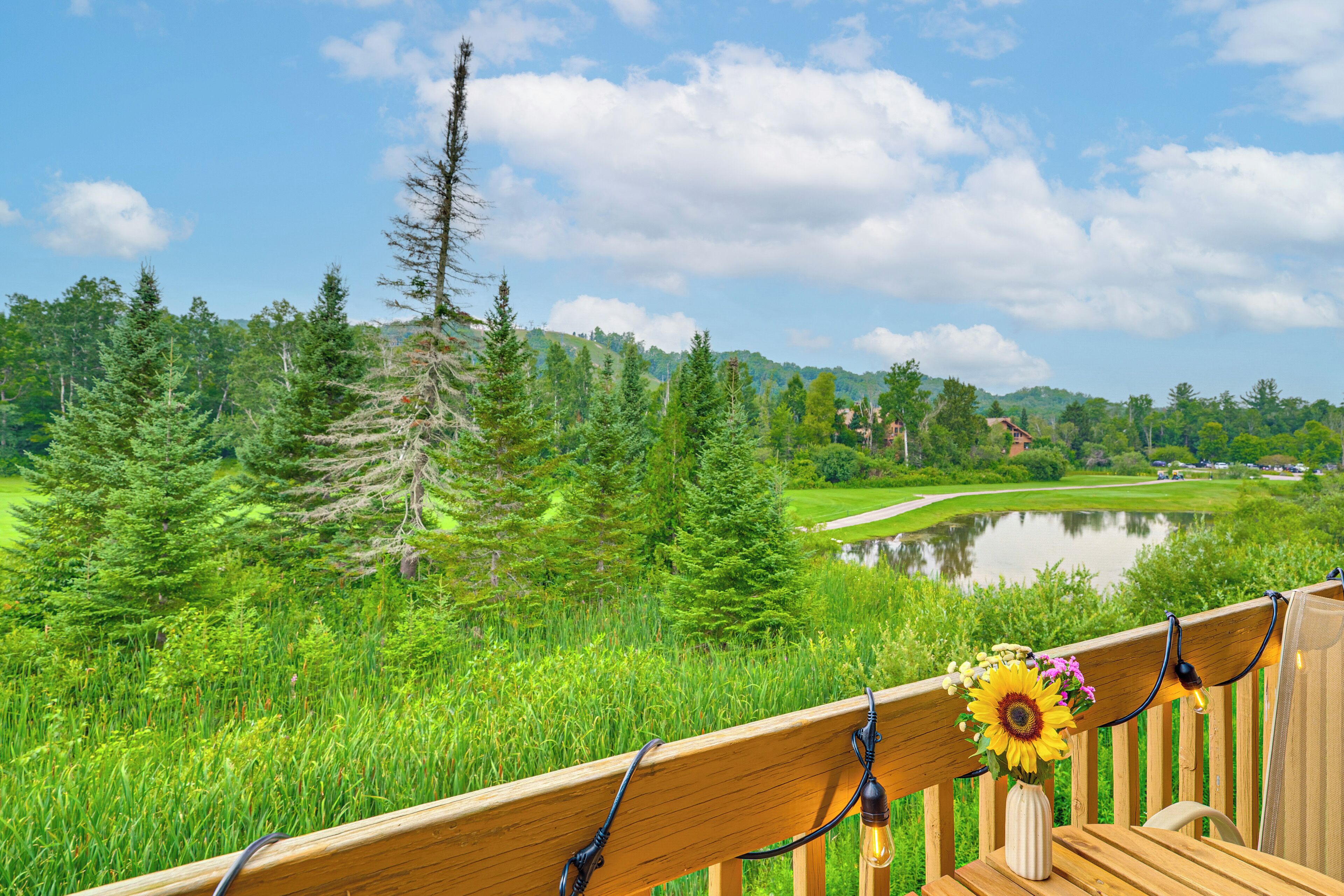 A wooden deck with a wooden railing, a vase with flowers, and a view of a golf course and lake.