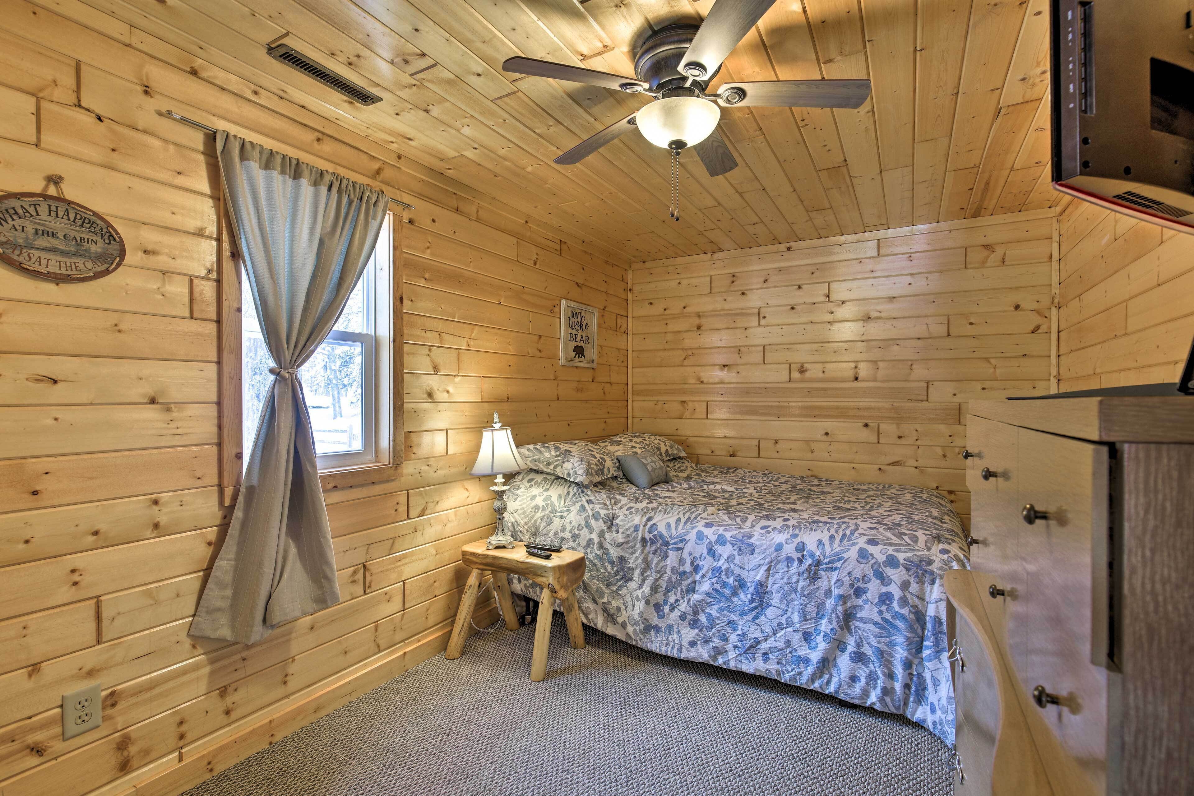 Una habitación de cabaña de madera con cama, ventilador de techo, ventana con cortinas y una mesita con lámpara.