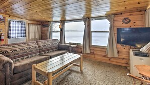 A cabin interior with a wooden ceiling, a sofa with a patterned cover, a small table, a television, and a view of a lake through large windows. - Charming Lost Lake Cabin: Fish, Boat & Hunt! (St Germain)