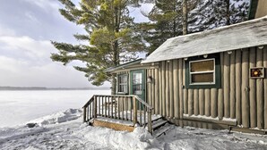 A log cabin with a wooden porch and a window, surrounded by snow and trees. - Charming Lost Lake Cabin: Fish, Boat & Hunt! (St Germain)
