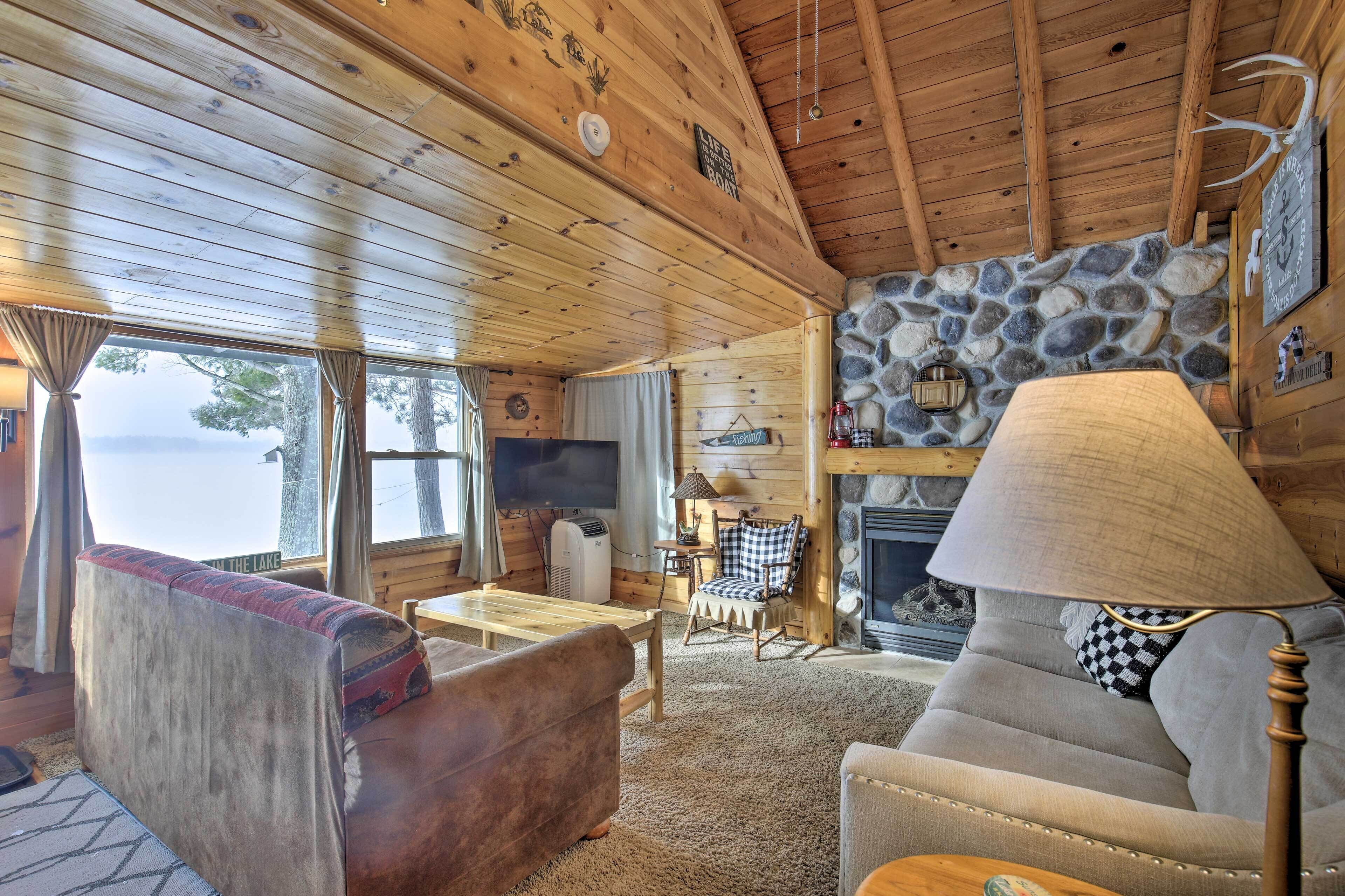 Een gezellig cabin interieur met een stenen openhaard, houten plafondbalken, een comfortabele zithoek en een groot raam met uitzicht op een meer.