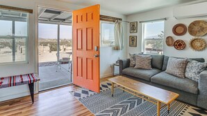 A living room with a grey sofa, wooden coffee table, and a bench. There are woven baskets on the wall and a door leading to a balcony. - Boho Bungalow w/ Hot Tub: 16 Mi to Joshua Tree NP (Twentynine Palms)