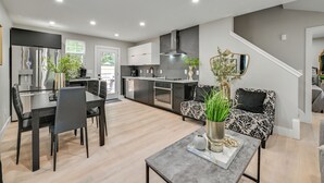 Una cocina moderna con electrodomésticos de acero inoxidable, una zona de comedor con una mesa y sillas negras, y una sala de estar con un sofá estampado y una mesa de centro.