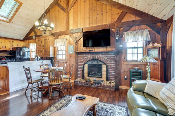 A rustic cabin interior with a fireplace, wooden furniture, and a television mounted above it. - Lakeside Log Cabin w/ Porch in Monadnock Region (Rindge)
