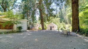 Une terrasse en gravier avec un espace repas, entourée de grands arbres et de deux petites cabanes.