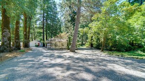 Une allée de gravier mène à un petit bâtiment blanc avec une porte rouge, entouré de grands arbres dans une zone boisée.