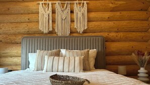 A cozy log cabin bedroom with a bed, macramé wall hangings, and a woven basket on the bed. - Sub Stele Chalets (Holbav)