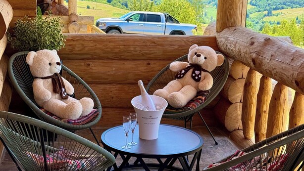 A wooden cabin with a balcony overlooking a scenic landscape. There are two wicker chairs with plush teddy bears, a small round table with a bottle of champagne, and a car parked outside. - Sub Stele Chalets (Holbav)