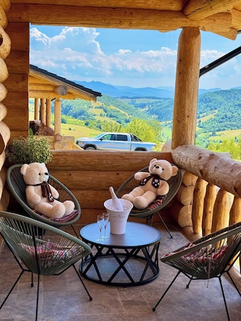 A wooden cabin with a balcony overlooking a scenic landscape. There are two wicker chairs with plush teddy bears, a small round table with a bottle of champagne, and a car parked outside.