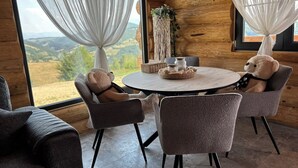 A cozy cabin interior with a round table, four chairs, and a view of the outdoors through large windows with sheer curtains. - Sub Stele Chalets (Holbav)