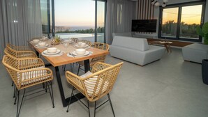 A modern dining area with a large table set for six, surrounded by wicker chairs, a sofa, and a TV. - Antiope Retreat (Pitsidia)