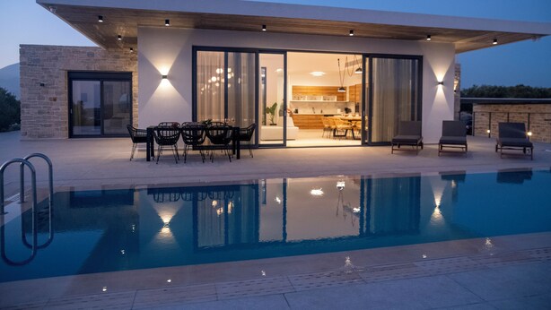A modern outdoor pool area with a dining setup and a kitchen visible through glass doors. - Antiope Retreat (Pitsidia)