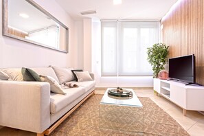 A modern living room with a sofa, a glass coffee table, a TV on a white cabinet, and a large window with blinds. - Luminous 1 Bedroom Apartment in the Heart of Seville. Murillo V (Seville)