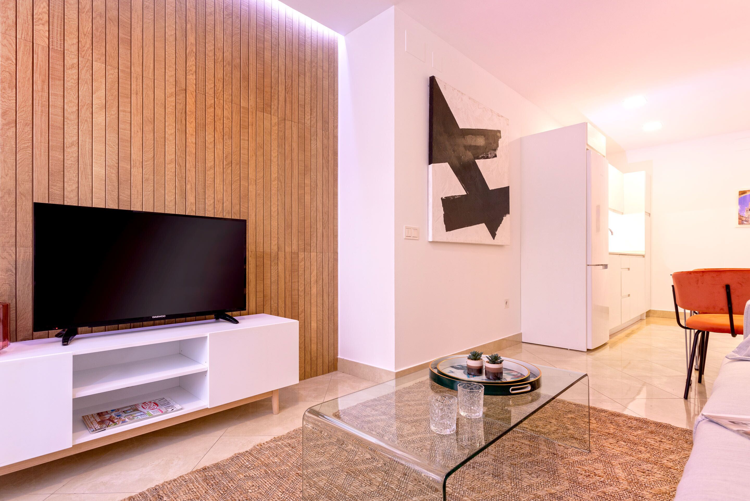 A modern living room with a flat-screen TV on a white console, a glass coffee table, and a large abstract painting on the wall.