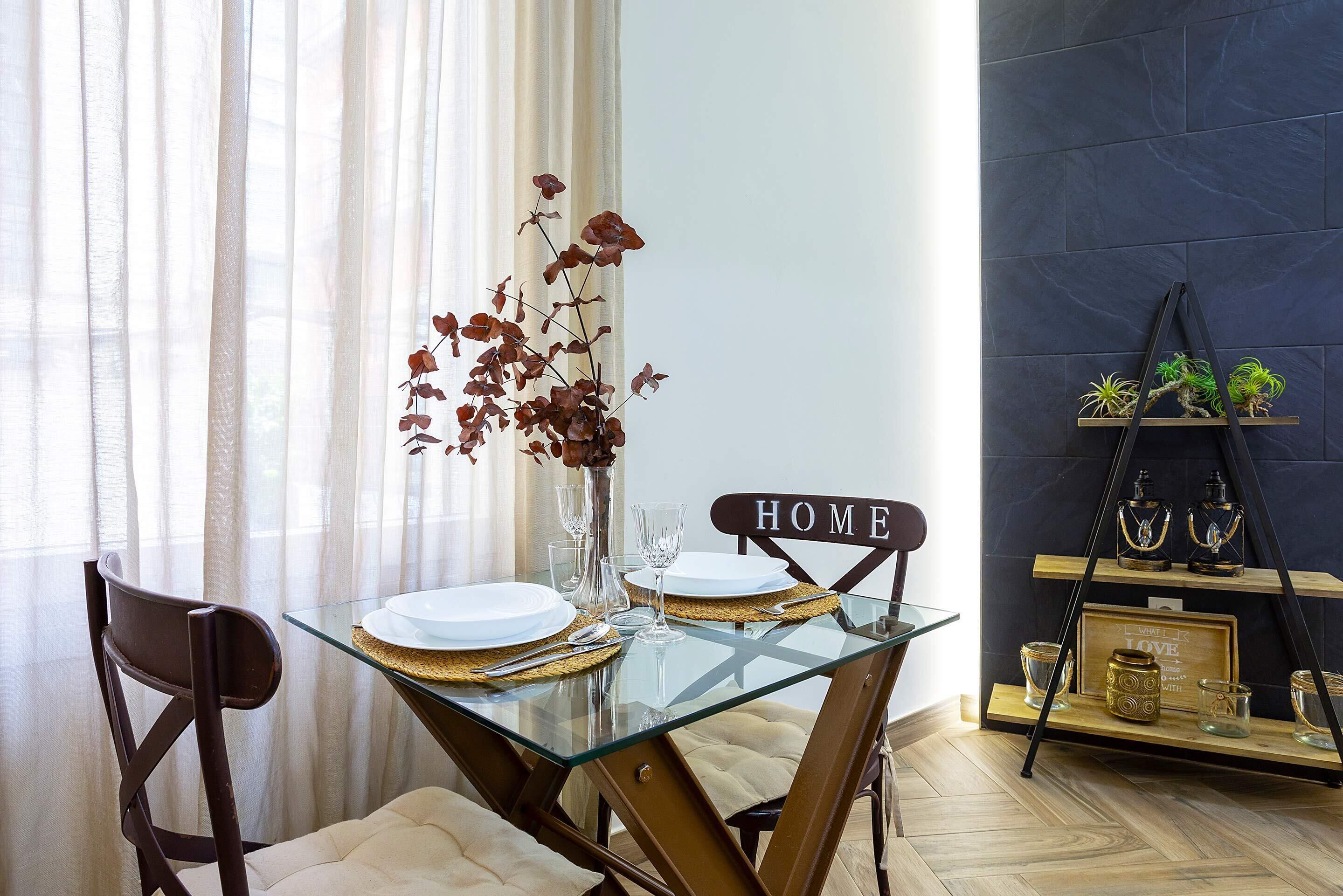Une table à manger avec un plateau en verre et des assiettes à bordure dorée, un vase contenant des feuilles séchées et une chaise ornée d’une inscription « HOME ».