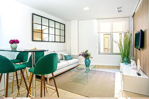 A modern living room with a glass coffee table, green chairs, a white sofa, and a TV mounted on the wall.