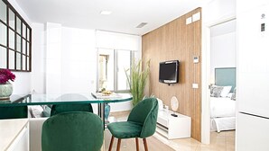 A modern hotel room with a glass dining table, two green chairs, a flat-screen TV mounted on a wooden wall, a bed with white bedding, and a large window. - Magnificent Apartment With Perfect Location. Murillo III (Seville)