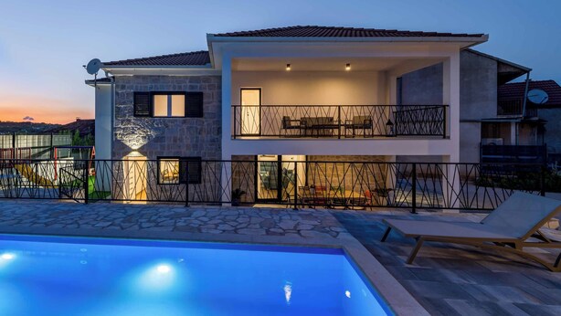 A modern two-story house with a swimming pool, stone facade, and a balcony with outdoor furniture.