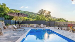 Ein Swimmingpool mit Liegestühlen und Sonnenschirm, umgeben von einer Steinmauer und Blick auf einen bewaldeten Hügel.