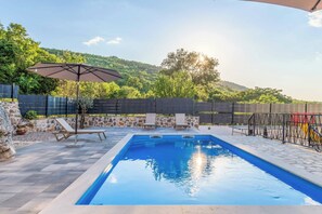 Piscina com espreguiçadeiras e um guarda-sol, cercada por um muro de pedra e vista para uma colina arborizada.