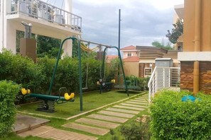 A playground with swings and a slide, surrounded by greenery and a building. - David & Jens 's Suite in Camella Manors Verdant (Puerto Princesa)