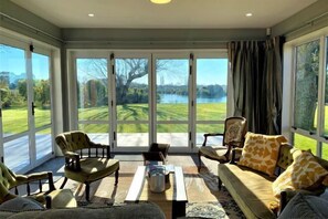 A room with a large window, a sofa, chairs, a coffee table, and a view of a lake and greenery. - Calm Swan Villa (Christchurch)