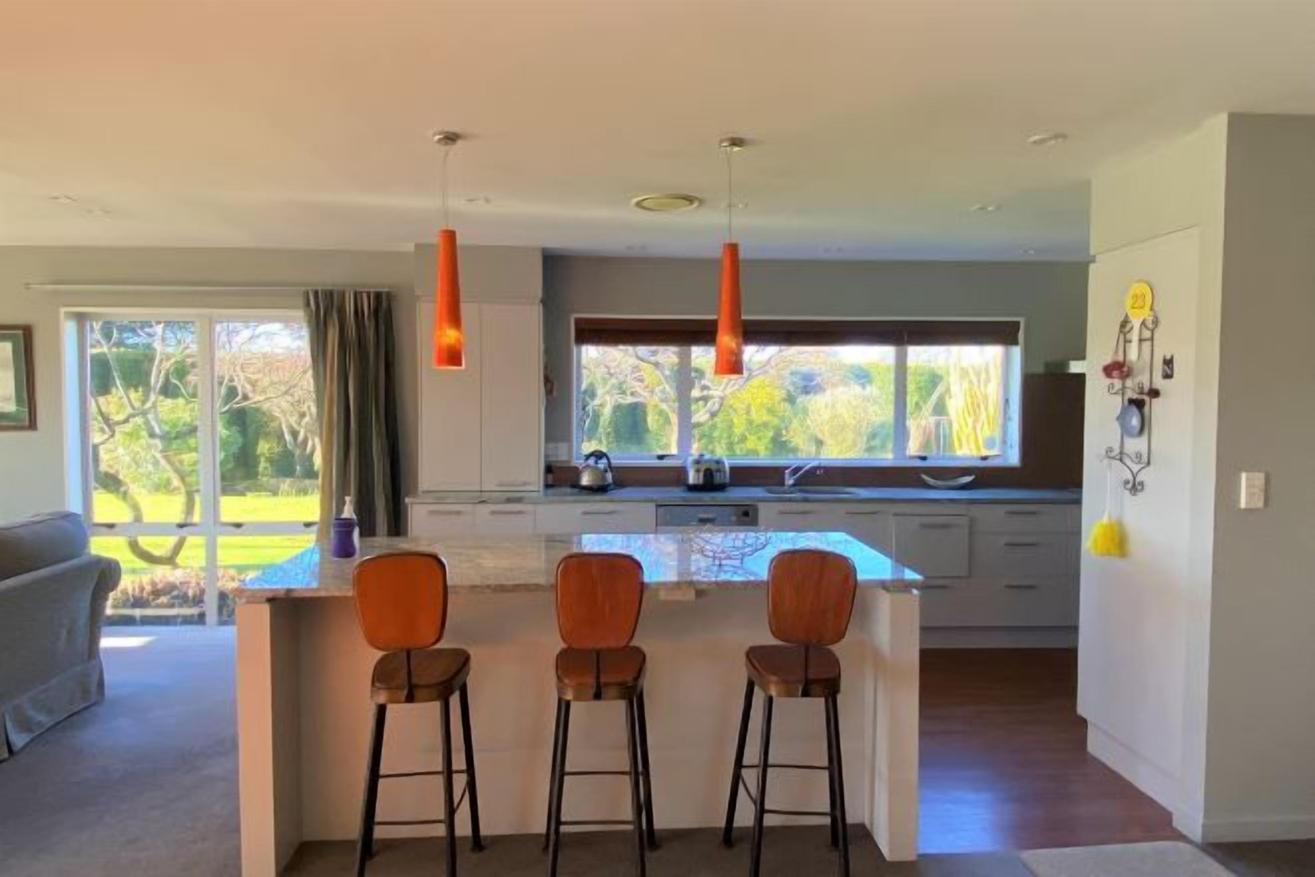 Una cocina moderna con una isla central, taburetes de bar, luces colgantes y vistas al exterior.