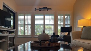 A living room with a television, a coffee table with decorative items, a sofa, and a window with blinds.