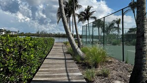 A wooden boardwalk beside a body of water, lined with palm trees and a chain-link fence.