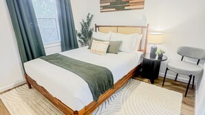 Un dormitorio con una cama, ventilador de techo, ventana con cortinas, mesita de noche con lámpara y una silla.
