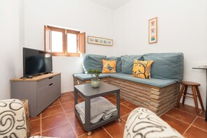 Ein Wohnzimmer mit einer Couch, einem Fernseher auf einem Schrank, einem kleinen Tisch mit einer Pflanze und einem Fenster.