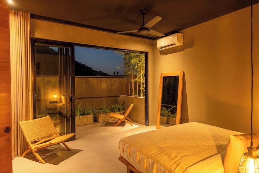 A bedroom with a bed, a chair, a balcony with plants, and a ceiling fan.