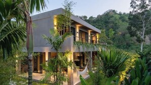 Een modern huis met twee verdiepingen en een plat dak, omringd door weelderige begroeiing en een hangmat.
