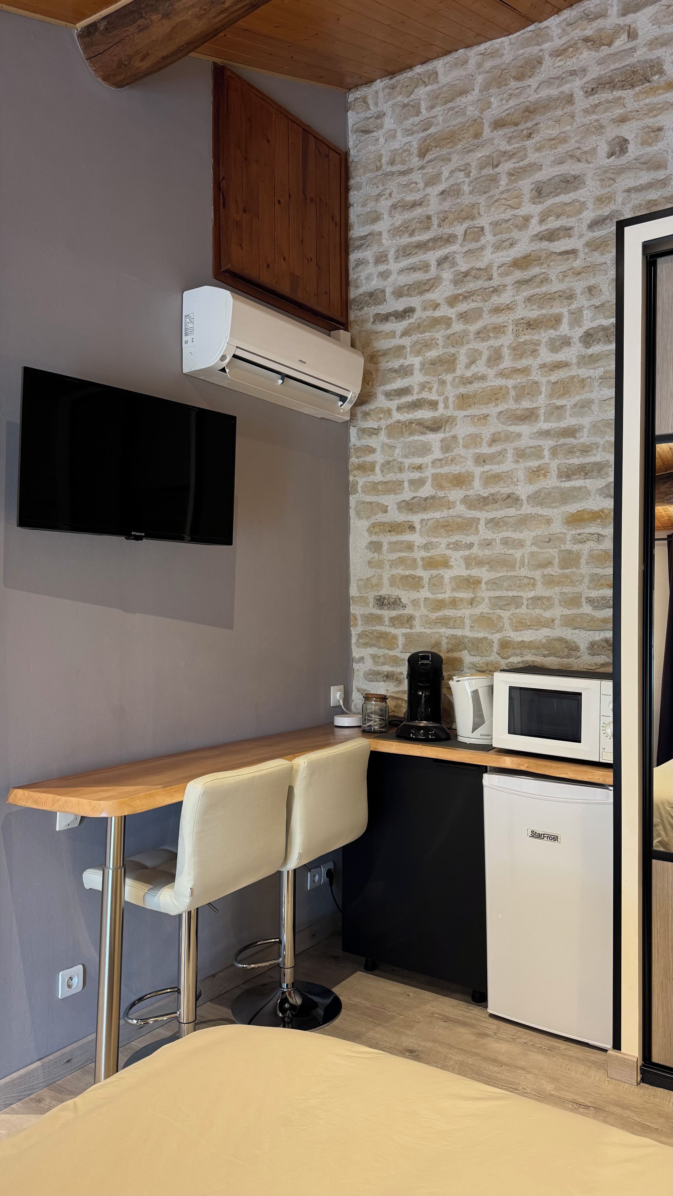 Een compacte keuken met een ingebouwde kookplaat, magnetron en koelkast. Er is een houten tafel met een stoel, en een flatscreen televisie aan de muur bevestigd.
