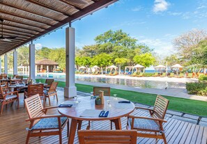 An outdoor patio with wooden tables and chairs, a pool, and umbrellas. - Ground Floor Condo in Bougainvillea 7103, Reserva Conchal Golf & Garden Views (Cabo Velas District)