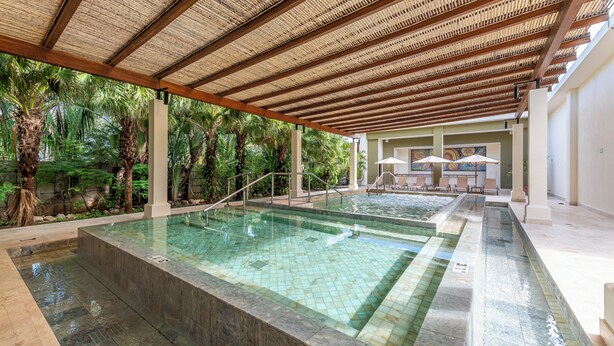 Un área de piscina con suelo de baldosas, una pérgola y palmeras.