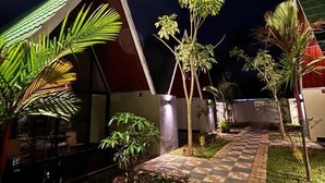 Vue nocturne d’un bâtiment moderne avec un chemin à motifs géométriques, des palmiers et un espace extérieur aménagé avec des sièges.