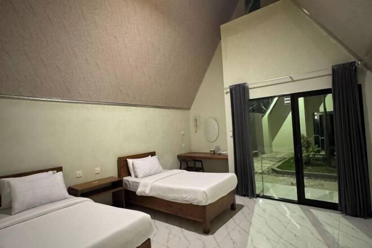 A hotel room with two beds, a desk, a chair, a small table, a mirror, and a sliding glass door leading to a balcony.
