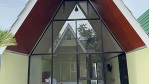 Un bâtiment de forme triangulaire doté d’une façade en verre, dont l’intérieur est visible et où l’on aperçoit un lit et une chaise, et qui est surmonté d’un toit végétalisé.