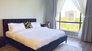 A modern bedroom with a large bed, a desk, and a view of greenery through the window.