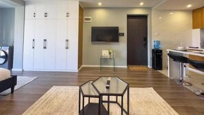A modern living room with a glass-top coffee table, a sofa, and a TV mounted on the wall. There is a door leading to another room and a small side table with a plant.