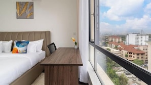 Quarto de hotel com uma cama, uma escrivaninha de madeira, uma janela com vista para a cidade e um quadro na parede.